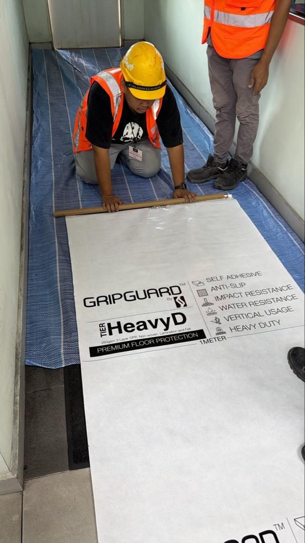 A worker in a hard hat and safety vest unrolling GripGuard HeavyD, printed with the GripGuard logo and its features, onto a site floor