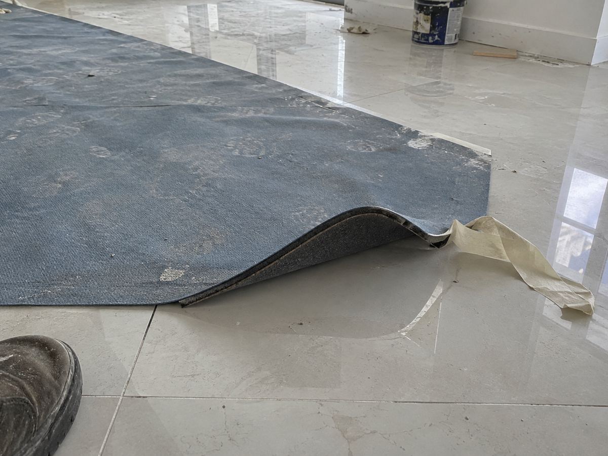 A loose floor protection mat on a polished tile floor with one corner curled up and the tape peeled off